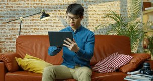 Joyful Asian young man sitting at red sofa with smile on face and browsing on tablet at home. Handsome male searching internet on device while resting on couch indoors. Leisure concept