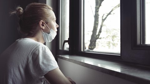 4k side view closeup of worried young Caucasian woman with blonde hair and white shirt wearing medical protection face mask watching the city by the window quarantine concept