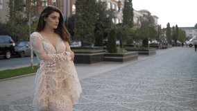 attractive model in long white tight dress with lacy bell sleeves stands on grey city paved road against trees slow motion