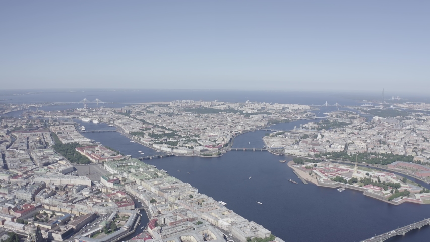 Panoramic aerial view of the city center of St. Petersburg reveals the Neva River cutting through the UNESCO World Heritage historic center, with multiple bridges spanning the wide waterway and connecting the dense urban fabric of baroque and neoclassical architecture. The camera captures the city from a high aerial perspective, showing the planned 18th-century layout with grand embankments, palace complexes, and the distinctive skyline that earned the city its nickname "Venice of the North." Founded by Peter the Great in 1703, the historic center preserves its original urban design with unobstructed sightlines across the river, where granite embankments and landmark structures line both shores. The scene unfolds under clear sunny skies in summer, with deep blue water contrasting against the pale stone and pastel facades of the historic buildings. Small boats move along the river while traffic flows across the bridges, conveying the scale and enduring vitality of this imperial Russian capital. The aerial perspective emphasizes the harmonious integration of water, architecture, and urban planning that defines this celebrated cultural destination. D-Log.