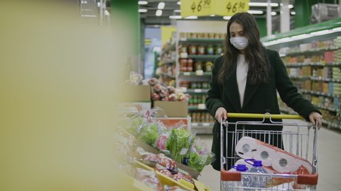 Young woman in protective medical mask buys food and hygiene items at supermarket during covid-19 coronavirus epidemic. Woman stocks up on food and toilet paper during quarantine and self-isolation