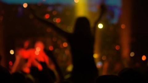 Super slow motion: people crowd silhouettes partying, cheering and raising hands up at rock concert in front of stage of nightclub. Bright colorful stage lighting. Nightlife and entertainment concept