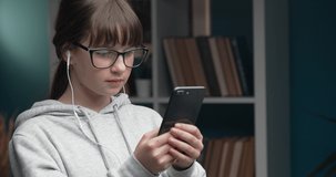 Attractive female teenager in eyewear sitting on sofa and using smartphone with earphones. Young girl with brown hair holding digital device at home.