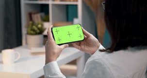View from back of woman with brown hair holding smartphone with chroma key green screen while relaxing on couch. Young lady looking at blank screen of digital gadget at home
