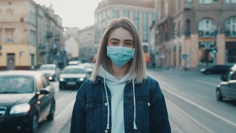 Young caucasian woman in medical mask standing in city street looking to camera road with cars on background Concept of health and safety life COVID-19 coronavirus virus protection pandemic in world