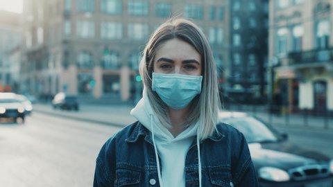Young serously woman in medical mask standing in city street looking to camera road with cars on background Concept of health and safety life COVID-19 coronavirus virus protection pandemic in world