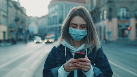 Young blonde woman in protective medical mask walks down to the street uses phone texts scrolls surfs the internet search news covid19 coronavirus virus protection pandemic city slow motion close up : vidéo de stock