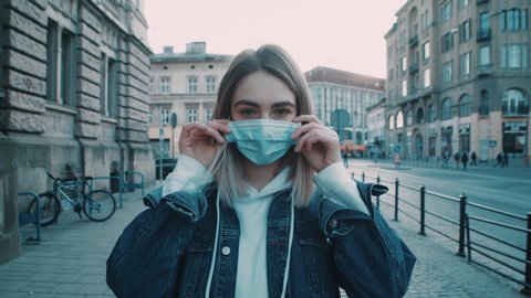 Pretty young caucasian woman stands in the city street puts on protective medical mask on face and looks to the camera covid19 coronavirus virus protection pandemic city slow motion close up outdoor