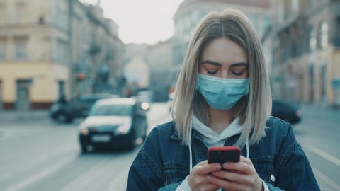Young blonde woman in protective medical mask stands in street and uses phone texting scrolling surfs the internet search news covid19 coronavirus virus protection pandemic city slow motion close up