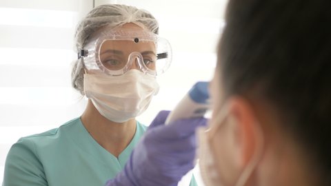 Nurse in a medical mask and glasses measures the patient's temperature with a non-contact infrared thermometer.