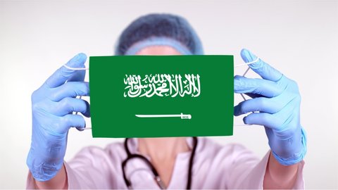 close-up. Doctor in glasses, blue medical cap, gloves holds in hands medical mask with Saudi Arabia flag. Physicians care, protection by state during coronavirus, global epidemic. Concept.