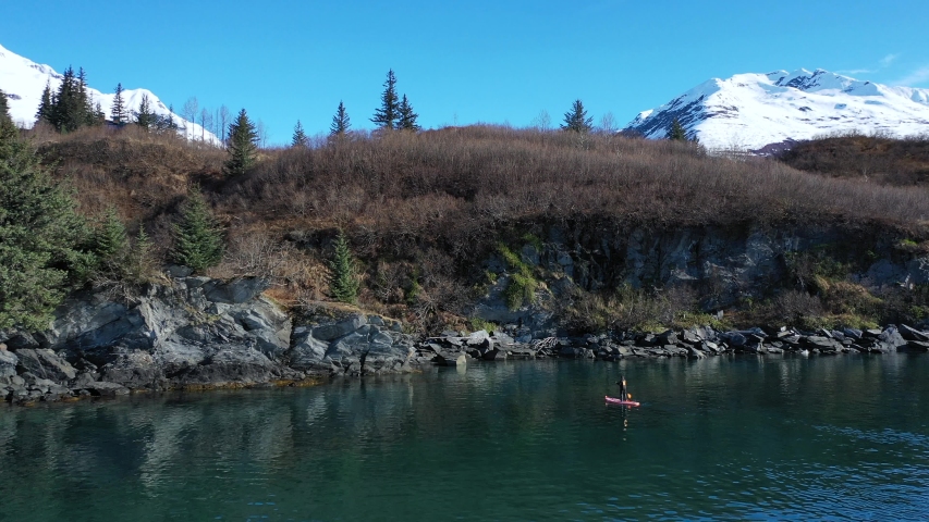 Boom up Drone shot of a paddle Boarder exploring the Alaskan Coast Line with Mountains in the background.