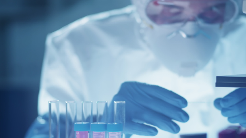 Rack focused shot of lab scientist in protective mask, suit, gloves and goggles placing glass slide in microscope and examining it while conducting research. Coronavirus pandemic concept