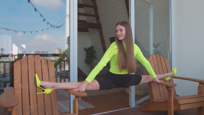 Lovely flexible young woman in stylsh high heels and leggings practicing side splits between two chairs on terrace in the morning. Graceful fitness female sitting on twine, doing stretching exercises. Royalty-Free Stock Footage #1049341378