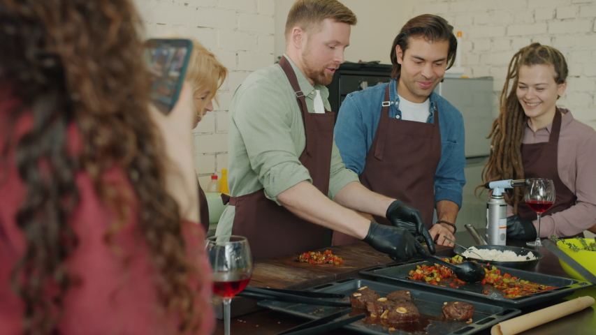 Group of cooks men and women making meal with fire while cooking class student in apron is taking pictures with smartphone camera in modern kitchen. Royalty-Free Stock Footage #1049349424