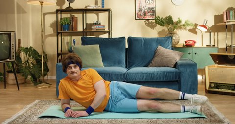 Dark-haired Caucasian athlete rising up his leg while exercising on yoga mat in living room. Funny sportsman of 50s in yellow t-shirt touching mustache while working out indoor. Training concept