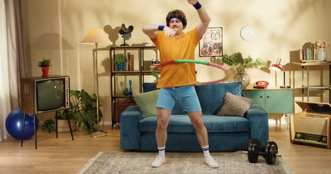 Funny Caucasian male fitness model with mustache fooling around while doing exercises with hula hoop near blue couch at home. Retro style amusing athlete working out with equipment indoors 60s concept