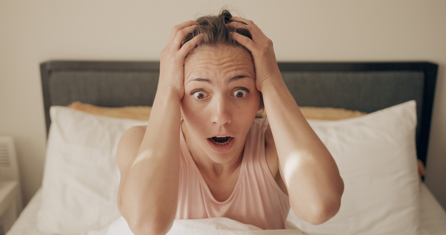 Front view of shocked young pretty woman oversleep and getting up fast while lying in bed . Beautiful girl waking up, holding head with hands and screaming while realising oversleep in morning