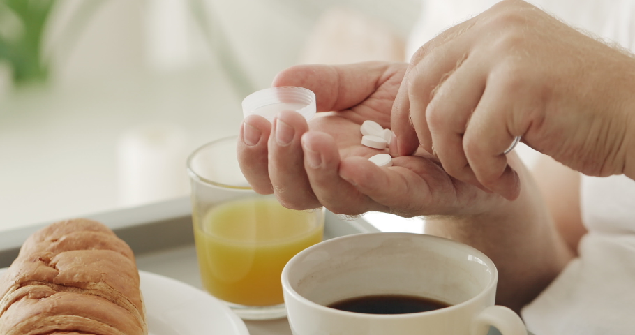 Close up view of man dropping pills from tablet bottle on palm hand and taking one to mouth. Man taking medecine while having breakfast in bed. Concept of health care