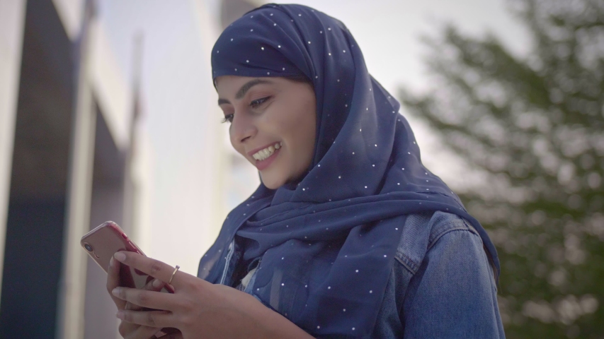A Muslim woman female standing outside commercial corporate complex wearing a Hijab headscarf using a smartphone mobile phone. A smiling happy Islamic office girl employee typing on a cellphone. | Shutterstock HD Video #1049360107