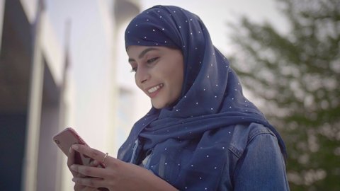 A Muslim woman female standing outside commercial corporate complex wearing a Hijab headscarf using a smartphone mobile phone. A smiling happy Islamic office girl employee typing on a cellphone.