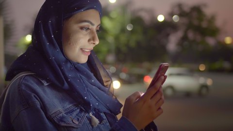 A muslim woman female standing outside outdoors wearing Hijab headscarf using smartphone mobile phone. A smiling happy Islamic girl typing on a cellphone against the moving traffic in the evening