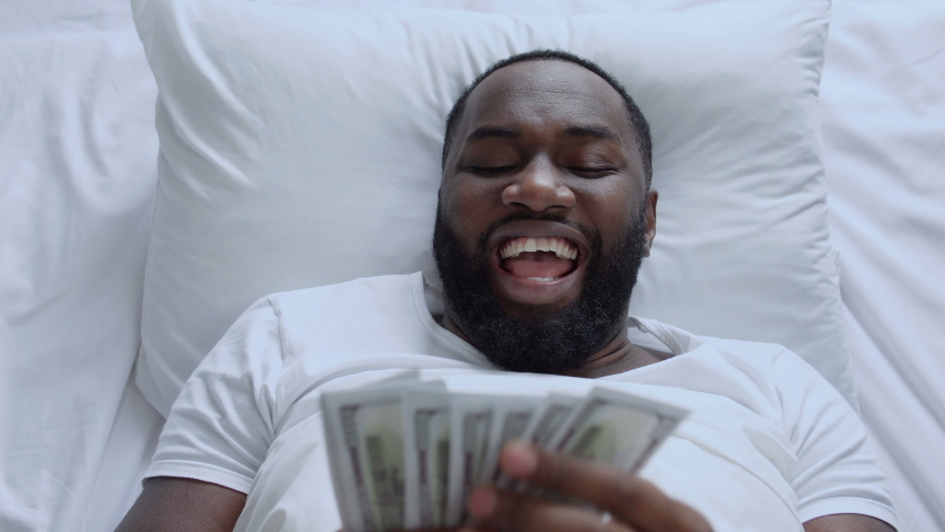 Cheerful afro-american man counting dollars and hiding under pillow lying in bed | Shutterstock HD Video #1049360332