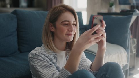 Rotation view happy young woman sitting on the floor near couch holding phone smiling enjoying using mobile apps for shopping having fun playing games chatting in social media at home slow motion