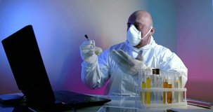 A man in a protective suit and a respirator looks at and discusses the substance in a test tube via a video link with a colleague