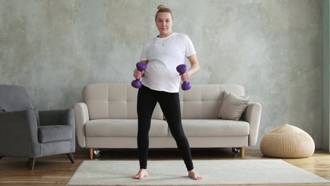 A pregnant blonde girl in a sports uniform laughs and dances with weights in her hands. Pregnant girl dancing at 9 months. Pregnant girl dancing