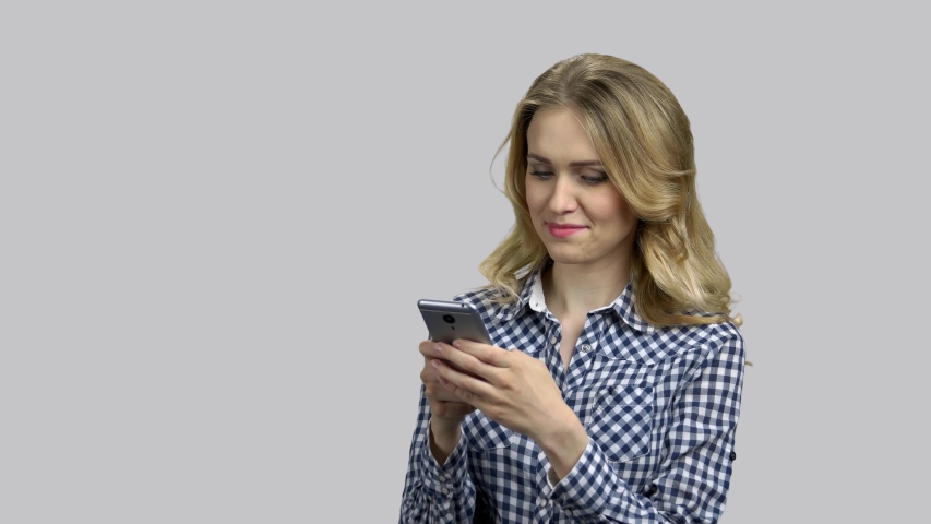 Young blond woman typing message on her smartphone. Attractive blond girl wearing casual shirt and chatting online, grey isolated background.