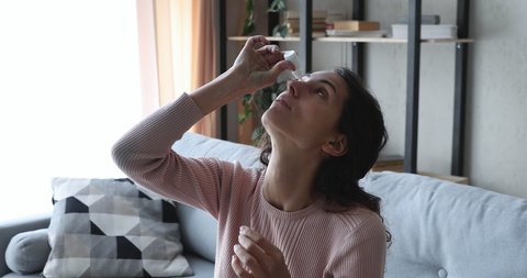 Young woman applying eye drops in dry irritated eyes sitting on sofa at home. Lady using eyedrops antihistamine ophthalmic antibiotic medication for allergy conjunctivitis infection treatment concept.