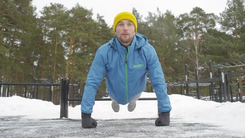 Slowmo shot of cheerful man in blue windbreaker and yellow beanie hat using low workout bar and doing push-ups on knuckles at outdoor gym in winter