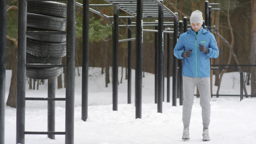 Medium shot with slowmo of active man in blue windbreaker warming up with jumping jacks and jogging in place while working out at outdoor gym in winter