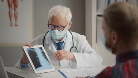 Doctor explaining lungs x-ray on tablet screen to young patient wearing facial mask to protect from contagious virus as covid-19 or flu. Practitioner showing x-ray with pneumonia symptom to patient