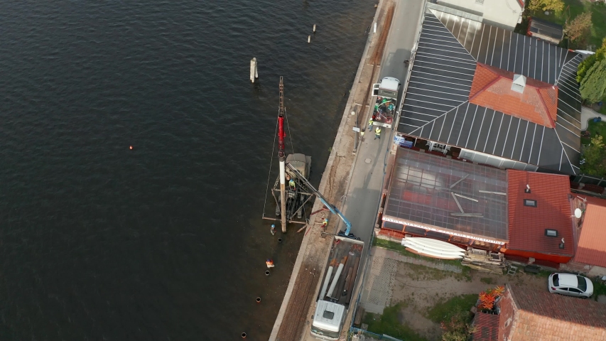 A construction site on the calm lake waters of Mikolajki, Poland with a tall drilling pipe pounding by the edge - Aerial shot