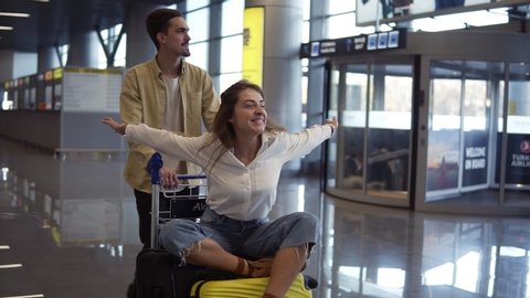 Romantic couple in airport. Attractive young woman and handsome man with suitcases are ready for traveling. Having fun on luggage trolley while waiting for departure. Woman immitating flying