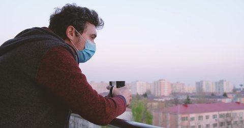 Man with medical mask leans on balcony railing holding a coffee mug, smells the coffee, and looks around in the distance. Isolation at home concept with space for graphics.