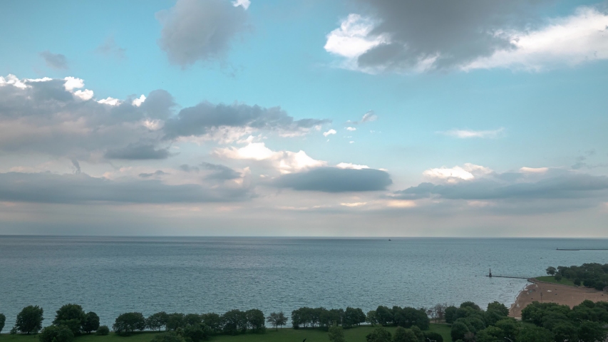 An aerial timelapse of fluffy white clouds filling a blue sky and then disappearing over Lake Michigan on the north side of Chicago and Foster Beach during sunset.