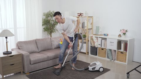 asian man with headphones is vacuuming living room and listening to online music streaming. chinese househusband cleaning house in a relaxed mood is humming and bobbing head with the upbeat song.