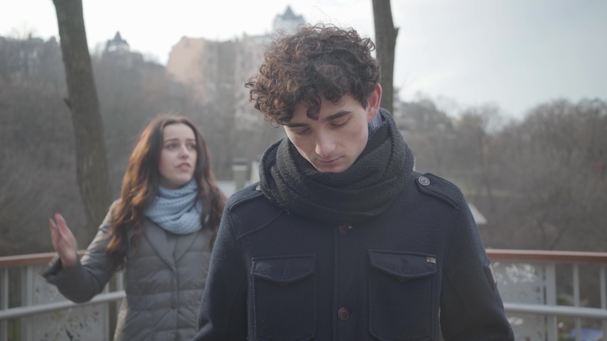 Portrait of young Caucasian man sighing and looking back at yelling girlfriend. Tired brunette boy exhausted from conflict with charming girl. Communication problems, conflict, jealousy.