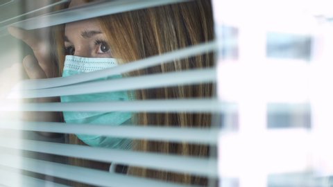 Sad young woman wearing medical protection mask looking out the window through the blinds at home. Home quarantine coronavirus. Social distancing, self isolation.