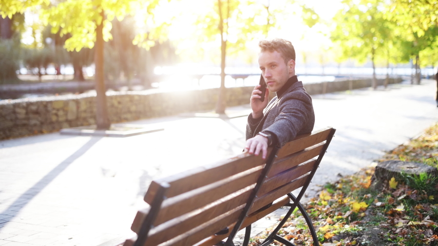Young man in formal clothes sitting in the autumn park at sunny day have a call.