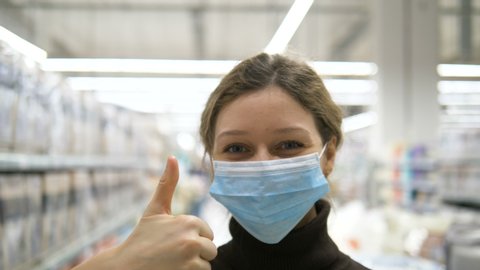 coronovirus protection. beautiful girl puts on a medical protective mask and gives thumbs up in supermarket. personal protective equipment