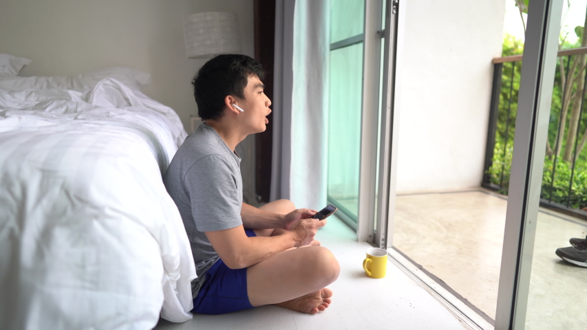 Young Asian man relax and chill sitting in the hotel room and drinking coffee also singing and listening a song.He is using the head phone.