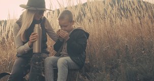 Happy mother and son hiking traveler camper drinking hot tea and relaxing in fall forest on sunny autumn day. Beautiful mountain landscape view. 4K slow motion video.
