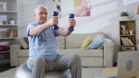 Elderly man sitting on exercise ball, watching online workout on laptop and training with dumbbells in the living room at home