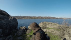 A male travel blogger comes to the river and rocks, takes selfie shots and videos of the landscape, background, in bright light, dressed in a brown jacket and backpack. gopro.