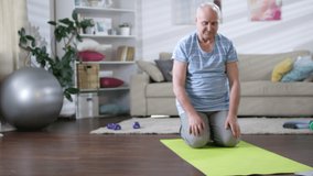 Senior man in sportswear doing upward facing dog exercise on mat in the living room with laptop on the floor while following online yoga workout