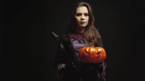 Young woman dressed up like a witch for halloween holding a scary pumpkin over black background